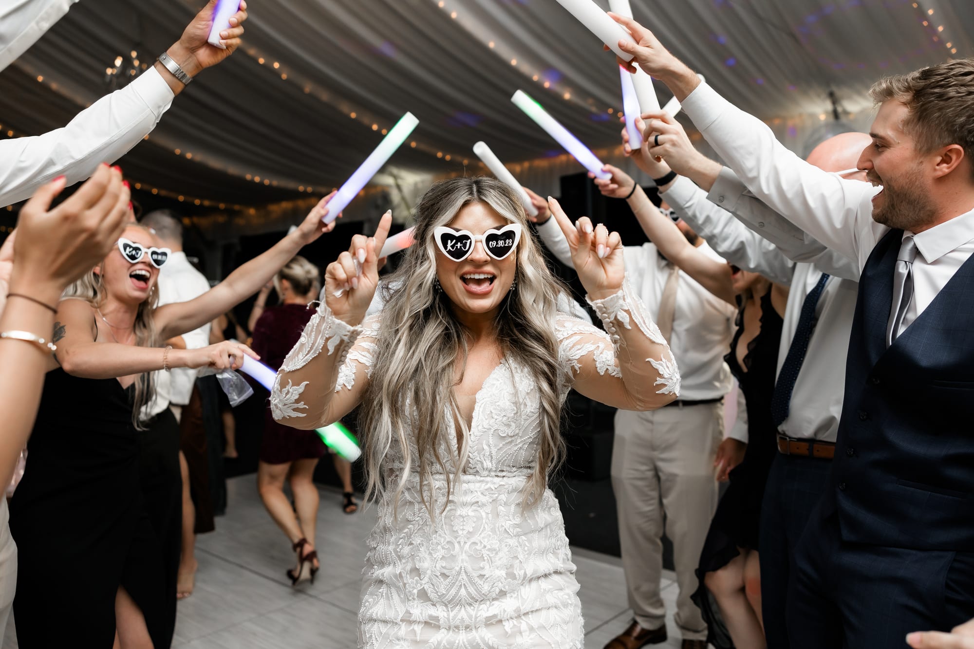 Bride in heart-shaped sunglasses leading a glow-stick exit from a draped tented reception