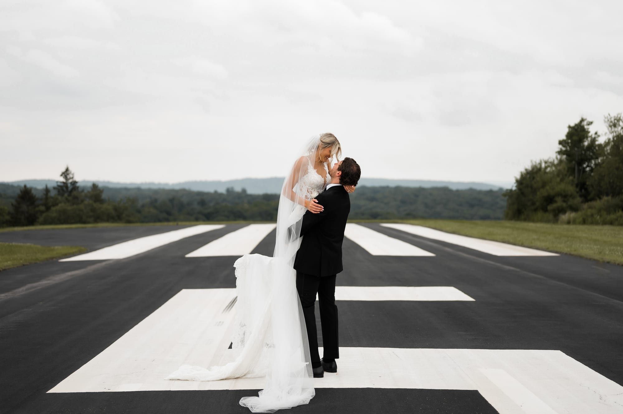 Bride and groom embracing on a private airstrip as her cathedral veil sweeps across the runway