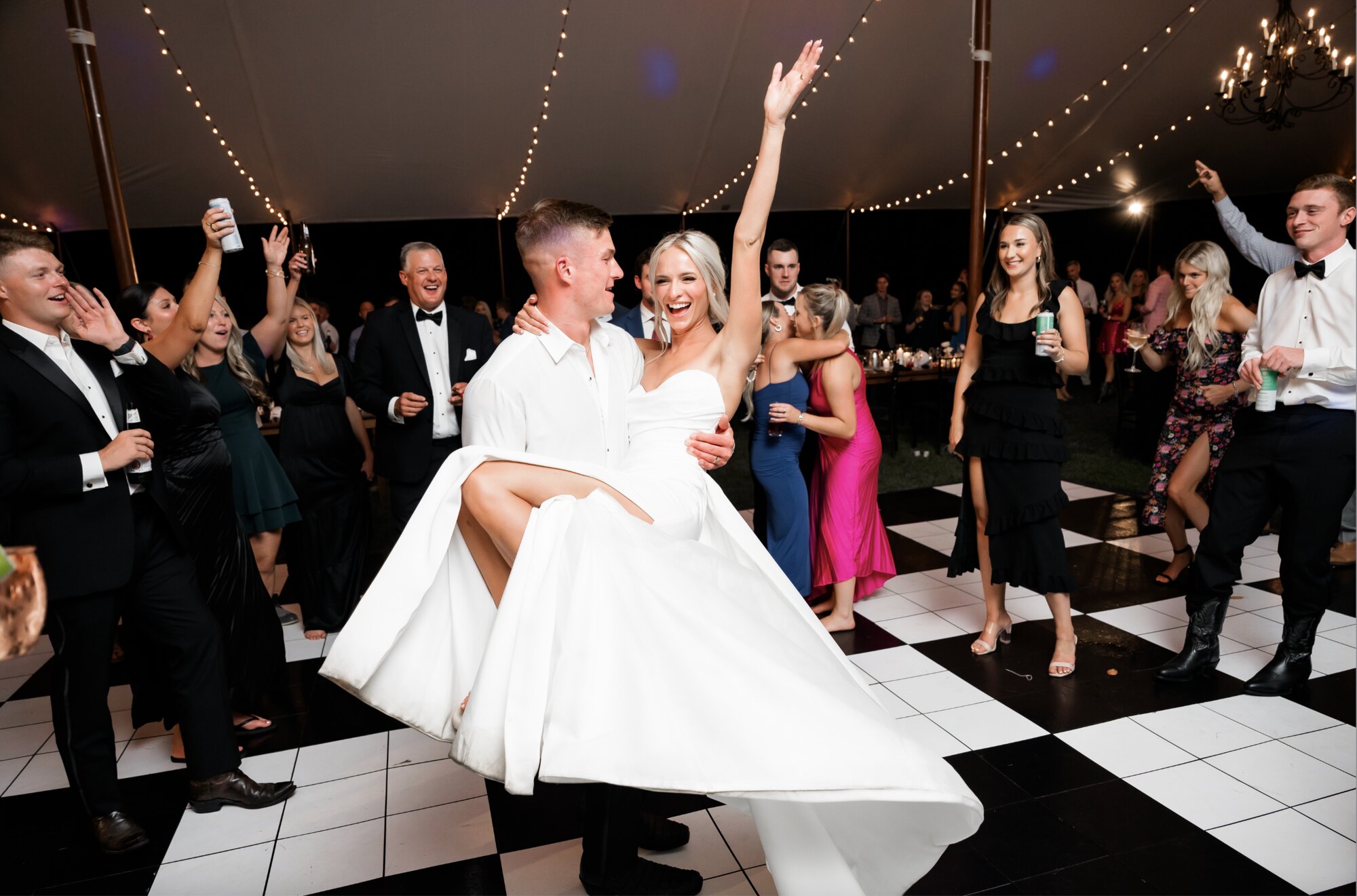 Groom lifting bride on a checkered tented dance floor as guests cheer